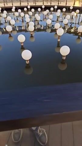 The video shows an outdoor scene at dusk. In the foreground, a dark body of water is filled with many round, white lights on poles, creating a reflective effect. A black railing runs along the edge of the water, and on top of the railing, a bottle of orange liquid is placed. Behind the water is a curved walkway with a wooden roof supported by poles and a metal railing. Several green, circular swings are visible on the walkway, and a few people are seated in them. The sky is a pale blue, indicating either early morning or late evening. In the background, there are some buildings and structures, including what appears to be a stadium light tower. The overall atmosphere is serene and modern, likely an outdoor entertainment or recreational area.