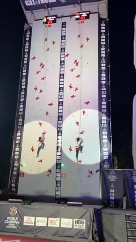 This video shows two athletes competing in a speed climbing event on a tall, artificial climbing wall. The wall is divided into two lanes, labeled A and B, each with a climber. Red climbing holds are scattered across the wall, and spotlights illuminate the climbers. Digital timers at the top of the wall display their progress, indicated by numbers. The athletes are wearing harnesses and are attached to ropes for safety. The background is dark, suggesting the event is taking place at night. Banners at the bottom of the wall indicate it is part of the World Climbing Championships in Chamonix. One climber, in lane A, is slightly ahead of the other, in lane B, and both are ascending the wall rapidly.