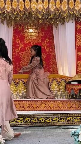 Two young girls in matching taupe-colored dresses are on a stage decorated with red and gold fabric. The girl on the left is standing and facing away from the camera, while the girl on the right is seated. The stage is adorned with ornate gold trim and hanging gold tassels, and the backdrop is a large red fabric with a gold pattern. There are white curtains on either side of the backdrop. In front of the stage, there are floral arrangements. The girl on the left appears to be offering something to the girl on the right, who is looking down. The scene seems to be an indoor setting, possibly for a performance or cultural event.