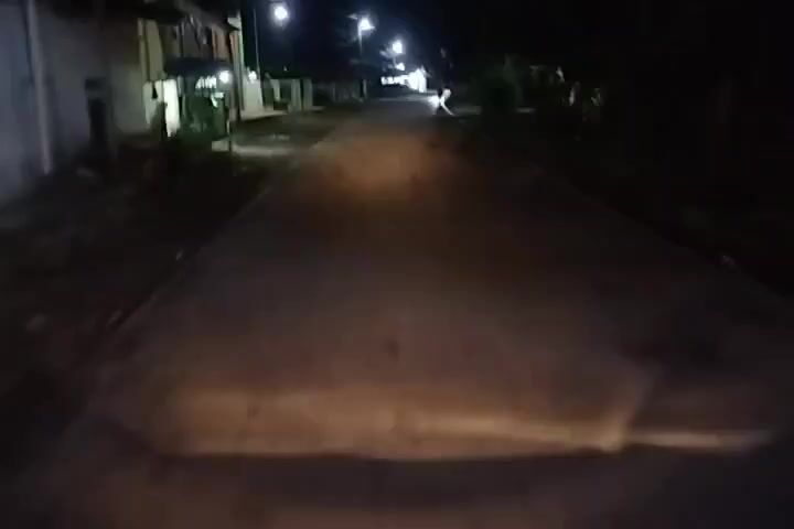 The video shows a first-person perspective of traveling down a paved road at night. The headlights of the vehicle illuminate the road ahead, creating a bright beam of light. On either side of the road, there are houses and vegetation, dimly visible in the darkness. Streetlights can be seen in the distance, casting a faint glow on the surroundings. The road appears to be empty except for the occasional approaching vehicle, indicated by bright headlights in the distance. The video captures the motion of traveling at a moderate speed, with the scene blurring slightly due to the movement.