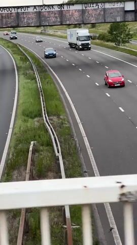 A high-angle shot shows a highway with multiple lanes of traffic. Several cars and a white truck are visible, moving in the same direction. The highway is bordered by green grass and trees. An overpass with graffiti is visible in the background. The sky is overcast. The camera appears to be positioned on a bridge overlooking the highway, with a white railing in the foreground.