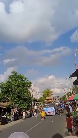 A parade proceeds along a road lined with onlookers. Several vehicles decorated with banners, flags, and objects on their roofs drive past. There are people dressed in costume in front of the first vehicle. The crowd is standing on both sides of the road. The sky is blue with some clouds. There are buildings, a power line, and a tree visible along the side of the road. The perspective is that of a person standing in the crowd watching the parade.