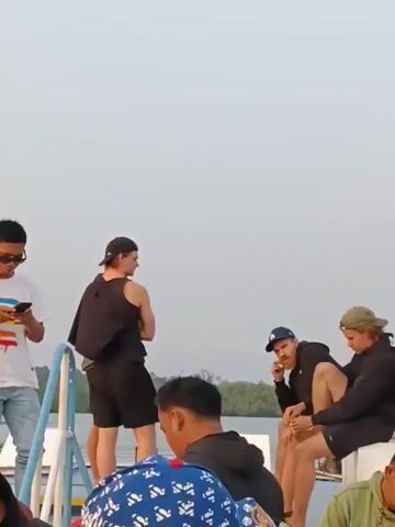 The video shows a group of people on what appears to be a ferry or boat. The sky is overcast and the water is calm. In the foreground, a man in a white t-shirt and jeans is looking at his phone. To his right, a man in a black tank top and shorts stands facing away from the camera, holding a jacket over his shoulder. To the right of him, two men are sitting on a ledge. The man on the left, wearing a blue cap, is on his phone. The man on the right, wearing a green cap, is looking down. Further in the background, on the shore, there is a small building with a red roof and some boats. The video pans slightly to the left, revealing more of the boat's railing and a man in a black t-shirt talking on his phone. The overall atmosphere appears relaxed, with people engaged in various activities.