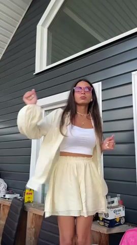 A young woman with long dark hair, wearing a white crop top, a yellow oversized button-down shirt, and yellow shorts, dances in front of a dark gray building with white trim. She has on pink sunglasses and jewelry. Her arms are moving and her mouth is open as she sings or talks. To the left of the woman on a wooden railing, there is a yellow bottle and a black chair. To the right, there is a box of Corona beer.