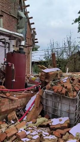 The video shows a partially destroyed brick building. The roof is significantly damaged, with exposed rafters and debris scattered around. Sections of the brick walls have collapsed, revealing interior spaces and equipment.  A large red tank sits amid the rubble, connected to a network of pipes and valves. Red plastic tubing is visible amongst the broken bricks and debris. A large plastic container, partially hidden by brick rubble, can also be seen. The scene suggests significant damage from an explosion or structural failure. The sky is overcast.