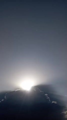 A video shows a sun peeking out from behind a cloud in a gray sky. The sun emits rays of light, creating a halo effect around the cloud. A faint crescent moon is visible in the upper portion of the sky. The video is brief and shows no people or other objects.