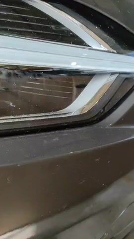 The video shows a close-up, low-angle shot of a car's headlight. The headlight is dark and appears to be either off or covered. The outer lens of the headlight has some dust and a few minor scratches. The chrome trim around the headlight is visible, as is a portion of the car's black bumper. The background is blurred and dark, suggesting an indoor environment like a garage or workshop. There are no people or actions depicted in the video.