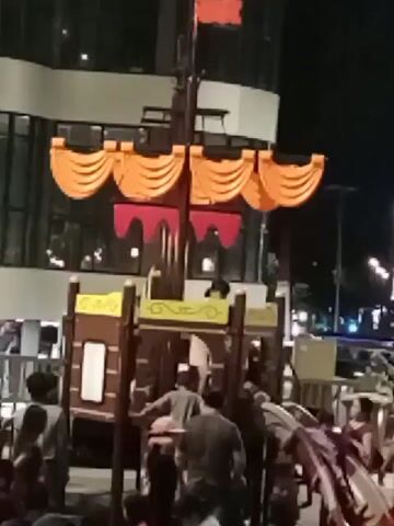 The video captures a lively scene at night, likely a public gathering or event. The foreground is filled with a crowd of people, their faces mostly obscured. In the background, there appears to be a temporary structure or stage decorated with orange and red banners, possibly for a festival or celebration. Behind this, a modern building with glass windows is visible, reflecting lights. There's an atmosphere of activity and movement, with people milling around and interacting. The lighting is predominantly artificial, suggesting it's after sunset.