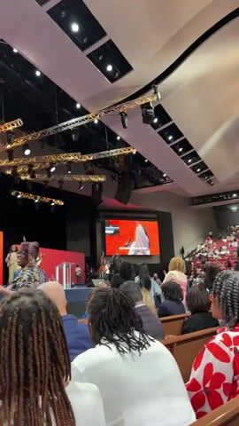 The video captures an indoor scene from a low angle, looking towards a stage in a large auditorium. The foreground shows the backs of several people with braided hairstyles, some wearing white and red patterned clothing. In the middle ground, a diverse crowd is seated in pews, facing a raised platform. On the platform, several individuals are visible, including a woman in a patterned dress speaking at a podium. Behind the speaker, a large screen displays an image of a person in a white garment. The background reveals a brightly lit stage with various lighting fixtures and architectural elements typical of a large venue or church. The overall atmosphere appears to be that of a formal gathering or service.