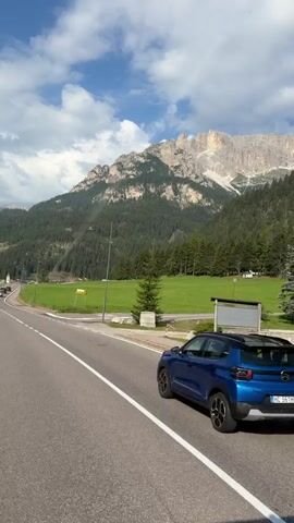 The video shows a road with a car driving on it. The road is bordered by a grassy area and trees. In the background, there are mountains. The sky is blue with some clouds. The car is dark blue and is driving in the right lane. There is another car in the distance. There are also some buildings in the distance.