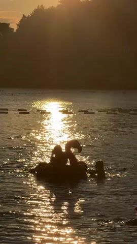 The video captures a serene lake scene during what appears to be sunset. The sun, low in the sky, casts a warm golden glow across the water, creating a shimmering reflection. In the foreground, silhouetted against the light, several individuals are enjoying a giant inflatable flamingo. They are actively playing and interacting in the water around the float. In the background, a line of buoys stretches across the lake, and the shoreline is lined with trees, also silhouetted by the setting sun. The overall atmosphere is peaceful and recreational.