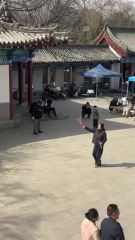 The video shows a stone paved square surrounded by traditional Chinese architecture. A woman is standing in the center, using a toy to create large soap bubbles. A small child runs towards her, trying to pop the bubbles with a toy sword. Other people are sitting or standing around the square, observing the scene. There are motorcycles parked near one of the buildings, and a couple of tents are set up on the right side of the square. The weather appears to be sunny.