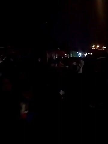 The video shows a dark outdoor scene with a crowd of people. There are some lights visible in the distance, suggesting an event or gathering. The individuals in the crowd are mostly obscured by darkness, but some appear to be wearing casual clothing. The atmosphere is lively, with people interacting. The setting appears to be a public space, possibly a street or a plaza, at night.