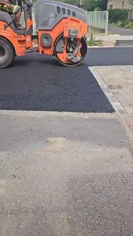 This video shows a road roller, an orange and grey heavy machinery vehicle, compacting a fresh layer of asphalt. A person, wearing an orange shirt and a hard hat, is operating the roller from the driver's seat. The roller has a large drum at the front and two large wheels at the back. The background suggests an outdoor setting, likely a street or road undergoing construction, with buildings and trees visible. The sun appears to be out, indicating daytime. The asphalt being compacted is dark and granular, contrasting with the lighter, textured paving stones on the right side of the frame. A concrete curb separates the asphalt from the paving stones. The text on the side of the roller reads "OZ-TRANS" and "HAMM," along with a website address and phone number, likely indicating the company performing the work. The overall scene depicts a construction or maintenance activity on a road.