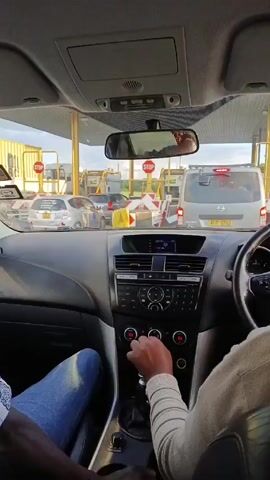 A static interior shot from the driver's perspective shows two people in a car, likely at a toll plaza. The driver, wearing a light brown shirt, is seen operating the gear shift. The passenger, wearing jeans and a patterned shirt, is seated to the driver's left. Outside the car, several other vehicles are waiting in line. Stop signs are visible above each toll booth. The car's dashboard features a stereo system and climate controls. The car's interior is primarily black and gray. The rear view mirror reflects the vehicles behind. The daylight suggests it is daytime.