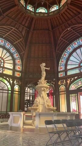 The video shows a wide shot of an indoor space with a dome-shaped wooden ceiling. In the center of the space is a white marble fountain with a statue of a nude female figure at the top. The fountain is surrounded by chairs. The walls of the space are adorned with stained glass windows in arched frames. The lighting is bright, creating a vibrant atmosphere. The video is shot from a low angle, looking up towards the ceiling.