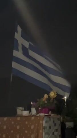 A Greek flag waves against a dark sky. Below the flag, on a table draped with a patterned cloth, a small arrangement of flowers and decorative figures is visible. The scene appears to be outdoors at night. In the foreground, a person's hand is holding a cigarette.