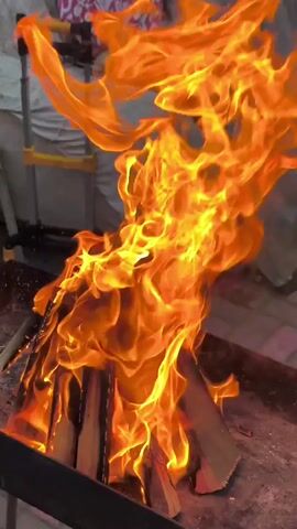 The video shows a close-up view of a bonfire with flames engulfing logs. In the background, a person wearing a patterned garment is partially visible, seated behind the fire. To the left of the fire, part of a metal stand with yellow handles is visible. The fire is the central focus, with flames reaching upwards and creating a dynamic visual. The overall environment appears to be outdoors.