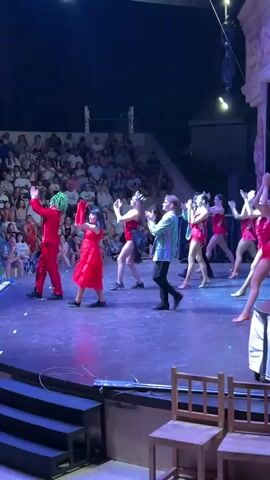 A group of performers are on a stage with a live audience in the background. The performers are dressed in costumes, with some wearing bright red outfits and others in yellow pants and white shirts. The performers are dancing and interacting with each other. The stage is lit with blue and purple lights, and there is a large audience seated in the background. There are also chairs in the foreground of the image.
