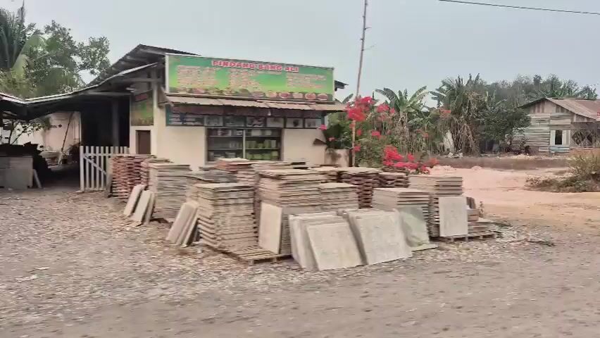 A slow-moving video captures a view from a vehicle traveling along a dusty road. On the left, a small shop with red flowers and stacked tiles is visible. Ahead, a dirt clearing is surrounded by lush banana trees and other vegetation. To the right, a collection of simple structures, including a dilapidated wooden building and a more modern concrete building, line the road. The sky is overcast. The camera continues to move, showing more of the roadside structures, some appearing to be small businesses or residences. There are various objects scattered around, including tires, barrels, and what appear to be tools or machinery. A red car is parked in front of one of the buildings. The overall impression is of a rural or semi-developed area with a laid-back, possibly commercial, atmosphere.