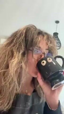 A woman with curly brown hair and glasses is drinking from a black mug with cartoonish eyes. She is wearing a dark apron with splotches of paint on it. The background is blurry but appears to be a kitchen, with a woven hanging light visible.