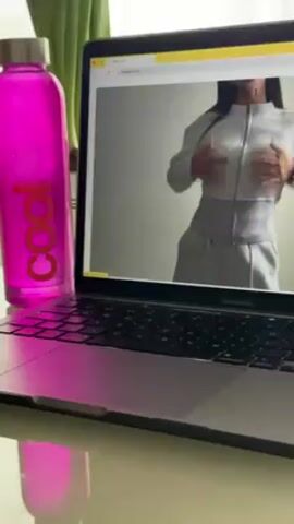 A MacBook Pro is on a desk, displaying a website with a model wearing a gray zip-up tracksuit. A bright pink water bottle labeled "cool" stands next to the laptop. A green plant is vaguely visible in the background.
