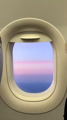 The video shows an intimate view from an airplane window, capturing a serene sky during what appears to be either sunrise or sunset. The sky displays a beautiful gradient, transitioning from a soft blue at the top to a vibrant band of pink and purple hues across the middle, then fading into a darker blue towards the bottom, suggesting the ocean or land far below. The window frame is a light, cream-colored plastic, typical of an aircraft interior. Part of a person's head or hair is faintly visible on the far right edge of the frame, indicating a passenger looking out. A small, dark speck, possibly a bird or another distant aircraft, can be seen in the blue part of the sky.

A female voice speaking Spanish is heard, likely an airline attendant making an announcement. The words, "que esta experiencia les deje buenos recuerdos" (that this experience leaves you with good memories), are clearly audible, suggesting a welcoming or farewell message. There is no visible text in the video. The overall mood conveyed is calm and picturesque.