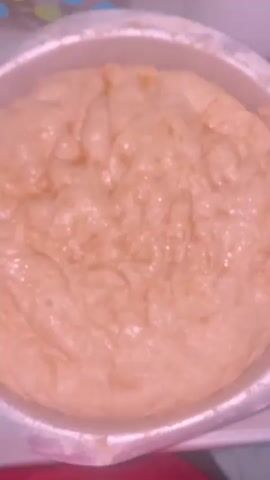 A close-up video shows a large metal bowl filled with dough. The dough is light brown and has a slightly oily sheen. The surface of the dough is uneven and appears to be rising, with some indentations and folds. The bowl itself is silver and has some flour dusted on its rim. The background is out of focus but appears to be a kitchen setting with a polka-dotted container and a white appliance visible. The video zooms in and out slightly, offering a detailed view of the dough's texture.