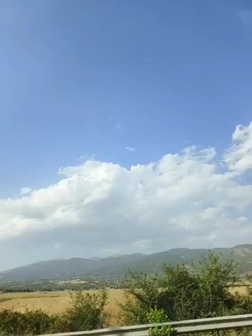 The video shows a wide shot of a rural landscape. A clear blue sky with scattered white clouds dominates the upper half of the frame. In the middle ground, a field of golden-yellow crops stretches across the width of the image. Behind the field, a range of hazy, green-covered mountains forms the backdrop. A small village or town is visible nestled at the foot of the mountains, with buildings and trees dotting the landscape. In the foreground, the edge of a dense patch of green foliage, possibly bushes or trees, can be seen. A tall, thin utility pole stands near the left side of the frame, cutting vertically through the middle ground and extending towards the sky. The overall impression is of a sunny day in a peaceful, natural setting.