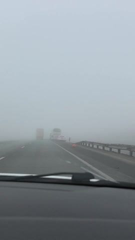 The video shows a view from inside a car traveling on a highway in heavy fog.  Several vehicles are visible ahead, including a yellow truck, a white van, a white sedan, and a large white passenger bus stopped on the shoulder of the road.  A person in dark clothing is standing behind the bus. A red warning triangle is placed on the shoulder behind the stopped vehicles. The windshield wipers of the car are visible. The fog is thick and obscures visibility significantly.