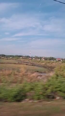 The video shows a blurry landscape from a moving vehicle. The sky is blue with scattered clouds. In the distance, there are houses with red roofs, vegetation, and a tall, white tower with red bands. The foreground is dominated by dried grass and some green foliage, also blurred due to motion. There's a dark horizontal object, possibly part of the vehicle, visible at the bottom. The scene appears to be a rural or semi-rural area.