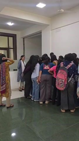 The video shows a group of young women gathered in a hallway with a green tiled floor. A woman in a white and blue patterned outfit is speaking to the group. Further down the hallway, a roll-up banner is displayed, featuring an image of a woman with elaborate makeup and the text "Solaah.in" and "ADHIRAAJ SOLAAH PE". The women are mostly facing the banner, and some are interacting with each other. The environment appears to be indoors, possibly a school or event venue, with white walls and a drop ceiling with lighting fixtures. The woman on the far left is wearing a yellow and red patterned dress.