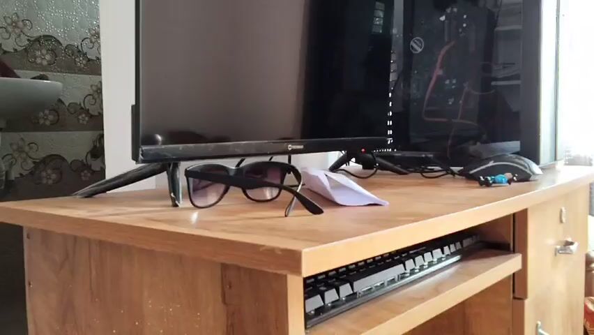 A computer monitor sits on a wooden desk in front of a computer tower. A pair of sunglasses and a folded piece of paper rest on the desk in front of the monitor. A keyboard is visible in a pull-out drawer below the desk.