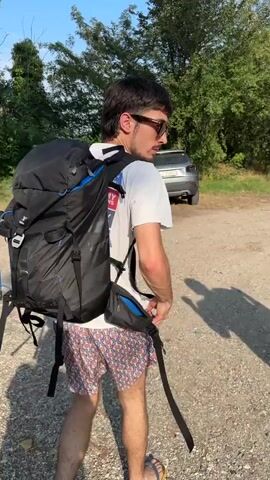 A young man with dark hair, wearing sunglasses and a white t-shirt with blue and red accents, along with colorful patterned shorts, stands facing away from the camera. He is wearing a large black backpack with blue accents. His right arm is extended forward, palm open, as if gesturing or directing someone. Beside him, a grey SUV is parked on a dirt path, surrounded by trees and vegetation under a clear blue sky. The perspective shifts to show another young man, this time wearing a dark t-shirt, light blue patterned shorts, a black baseball cap, and flip-flops. He carries a striped towel draped over his left shoulder. He is walking away from the camera, towards the parked SUV. Shadows of both individuals are cast on the ground, indicating bright sunlight.