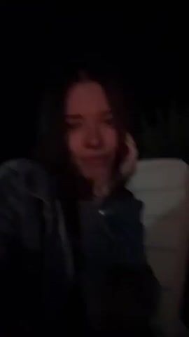 The video begins with a close-up of a young woman's face, illuminated by a dim light. She is looking down, with her hand resting on her cheek. The scene then shifts to a wider shot of a group of people gathered around a bonfire at night. Several individuals are seated on chairs or the ground, facing the fire. One person is playing an acoustic guitar. In the background, there is a large, white, tent-like structure and a white portable building. The fire is burning brightly, casting a warm glow on the scene. The atmosphere appears relaxed and convivial.
