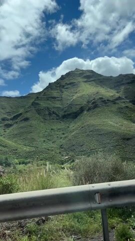 The video captures a scenic view of a mountain range under a sky with patchy clouds. The camera appears to be positioned from a moving vehicle, as indicated by the visible guardrail in the foreground. The mountains are covered in green vegetation, with a rugged terrain visible through the varying shades of green. The sky above is bright blue, punctuated by puffy white and grey clouds that add depth to the vista. The scene is well-lit, suggesting a clear day, and the focus is on the expansive mountain landscape.
