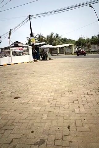 The video shows a street scene. In the foreground is a paved area with interlocking bricks, showing some dark stains. In the midground, there is a small roadside stall with a white roof and a white wall on the left. Two people are standing near the stall. A yellow truck is driving past the stall on the right side of the road. A person on a motorcycle is following the truck. In the background, there are trees and a fence. Overhead, there are power lines and a street lamp. The sky is clear and blue.