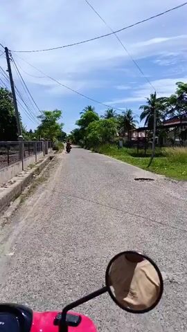 The video shows a first-person perspective of someone riding a scooter or motorcycle down a paved road. The road is lined with green vegetation, trees, and some houses. The sky is blue with scattered white clouds. There are other vehicles on the road, including at least two other motorcycles and a three-wheeled vehicle. The rider's reflection can be seen in the scooter's side mirror. The road appears to be in a rural or semi-rural area.