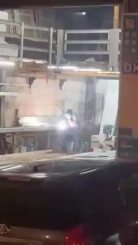The video shows a person welding inside a building at night. The individual is wearing protective gear and using a welding torch, producing bright sparks. The building appears to be a workshop or small industrial space, with metal materials and shelving visible in the background. The scene is viewed from outside, through what appears to be a car window.