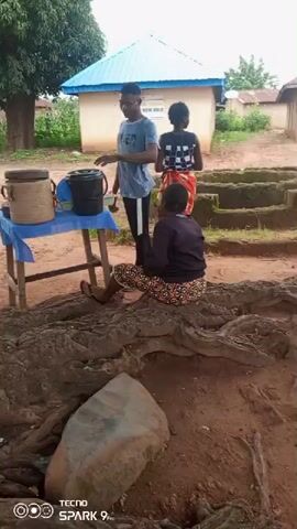 A video captures three people in an outdoor setting. A young man wearing a blue t-shirt and black pants stands behind a table covered with a blue cloth. On the table are two large brown containers and a black bucket. The man appears to be pouring something from a smaller container into the black bucket. A young woman, wearing a black and white patterned top and a red and white patterned skirt, stands next to him, looking at the bucket. In the foreground, a woman with braided hair, wearing a dark purple top and colorful patterned pants, sits on large exposed tree roots. She is looking towards the table. Behind the table, a building with a light blue roof and tan walls is visible. The ground is dirt, and there are some green plants and a large tree in the background. The video has a timestamp indicating it was recorded on a Tecno Spark 9 Pro.