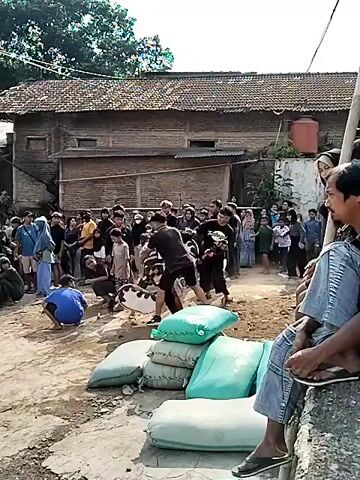 The video shows an outdoor gathering of people, with a performance taking place in the center. A group of individuals dressed in black are interacting with someone in elaborate, traditional attire, possibly a costume representing an animal or mythical creature. Other people, including children and adults, are observing the event. Some are standing, while others are sitting or crouching. The environment appears to be a village or rural area, with simple buildings in the background and sacks of what appear to be sand or grain piled in the foreground. The overall atmosphere seems to be one of community engagement and cultural celebration.