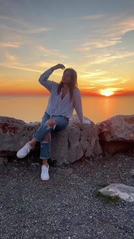 A young woman is sitting on a rock in front of a sunset over the ocean. She is wearing a striped blue and white shirt, ripped blue jeans, and white sneakers. She runs her hand through her hair and looks around. The sky is a mix of orange, pink, and yellow. The ground is covered in gravel. There are other large rocks surrounding her.