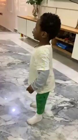 Here is a description of the video.

The video features a young child, likely a toddler, standing indoors. The child has dark, curly hair and is wearing a light-colored, long-sleeved shirt with small yellow flowers, green pants, and white socks. They are standing on a gray and white marble-patterned floor.  The child looks upwards initially, then turns their head and appears to be talking or singing. In the background is a white cabinet, a table with gold detailing, and a vase of flowers. The overall environment appears to be a modernly decorated living room.