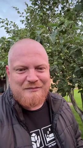 A bald, middle-aged Caucasian man with a reddish-brown beard is shown in a close-up shot. He is wearing a black puffer jacket over a black t-shirt with a graphic print. The man is standing outdoors, with green trees and foliage visible behind him. The sky appears overcast with hints of sunset colors. The man is smiling directly at the camera. The scene appears to be in a rural or suburban setting, possibly a backyard, with a wooden structure visible in the background. The camera is handheld and slightly shaky, suggesting it might be a selfie video or a casual recording. The overall atmosphere is informal and relaxed.