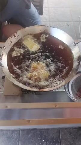 In the video, a large, tarnished, silver-colored metal pan filled with dark bubbling oil sits on a metal countertop, likely in a kitchen or food preparation area. Food is being deep-fried, as evidenced by the bubbling oil and the light-colored items submerged within it. A person wearing a blue shirt and pants is visible in the background, sitting or kneeling near the counter but not actively engaged with the pan. The setting appears to be indoors, with tiled flooring and a partial view of what looks like a sign or decoration. The camera remains static, providing a direct, high-angle shot of the cooking process.