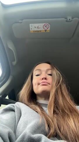 A young woman with long, light brown hair sits in a car, looking directly at the camera. She is wearing a light gray sweater and has her left hand resting on her head. Her face is made up, and she has visible dimples. She closes her eyes and then opens them, looking around the car before returning her gaze to the camera. Above her head, a sun visor is visible with an "AIRBAG" warning label written upside down. The interior of the car appears to be light gray or white.