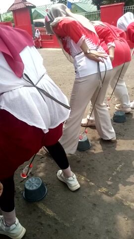 This media file features an outdoor scene at ground level, likely in Sukaraja, Indonesia, given the context. The primary subjects are several people, predominantly women, engaged in a game or competition. They are dressed in red and white attire, with some wearing hijabs. Their clothing suggests a coordinated event, possibly a school or community gathering.

The activity involves a relay race or a similar challenge where participants are tethered by strings to bottles, which are then being directed into buckets placed on the ground. The people are in a crouched or semi-crouched position, indicating exertion and focus on the task. The objects in the scene include plastic water bottles, gray buckets, and what appear to be red and white barriers or structures in the background, possibly part of the competition setup. The ground is asphalt, marked with shadows, suggesting it is daytime.

The overall mood seems to be one of energetic participation and possibly friendly competition. The lighting indicates it is daytime, with sunlight casting shadows. There are hints of other participants or spectators in the background, and some red banners or structures are visible. The image is captured from a low angle, emphasizing the participants and their actions. While no explicit text is clearly legible on the clothing or objects, the context points to a local event.