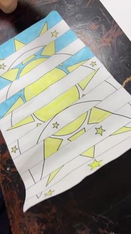 The camera focuses on a white piece of paper laid on a dark-colored table.  The paper is folded horizontally in an accordion style, creating several parallel strips.  These strips are colored with yellow and blue markers, and the design includes a sun-like shape with stars. The yellow marker is used to color the inner part of the sun and some stars, while the blue marker fills in parts of the background and sections of the sun's rays.  A white marker lies on the table near the paper, out of focus. A person's voice is heard in the background.
