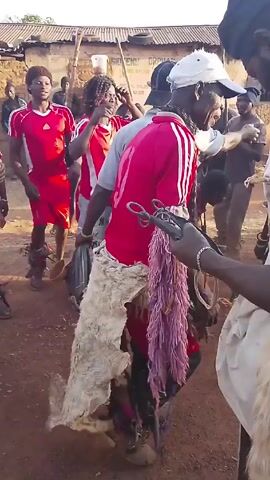 Several men are gathered in a dusty, outdoor area, some wearing red sports jerseys with white stripes. One man wears a white fur-like covering around his waist. Another man holds a collection of metal objects, possibly tools or decorations, tied together. The men are moving and talking, possibly dancing or participating in a cultural event. There is a building with the words "SIDEENU ORPHAN" visible in the background.