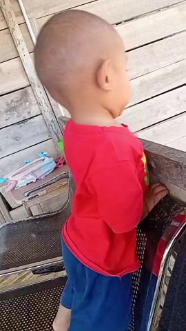 A toddler in a red t-shirt and blue pants is standing and holding onto the edge of a wooden structure, possibly a deck or porch. The toddler looks to the side with a surprised expression. In the background, there are wooden planks and a chair. The overall environment appears to be outdoors.